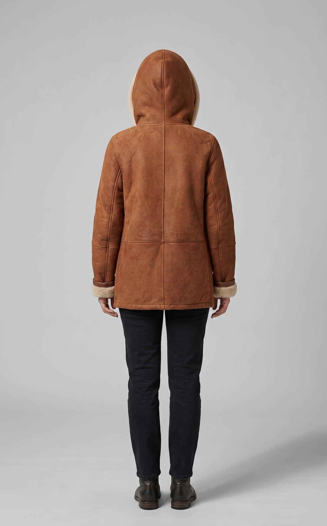 A person shown from the back wearing a brown suede-like hooded jacket with light beige trim, paired with dark pants and boots, against a plain light gray studio background.